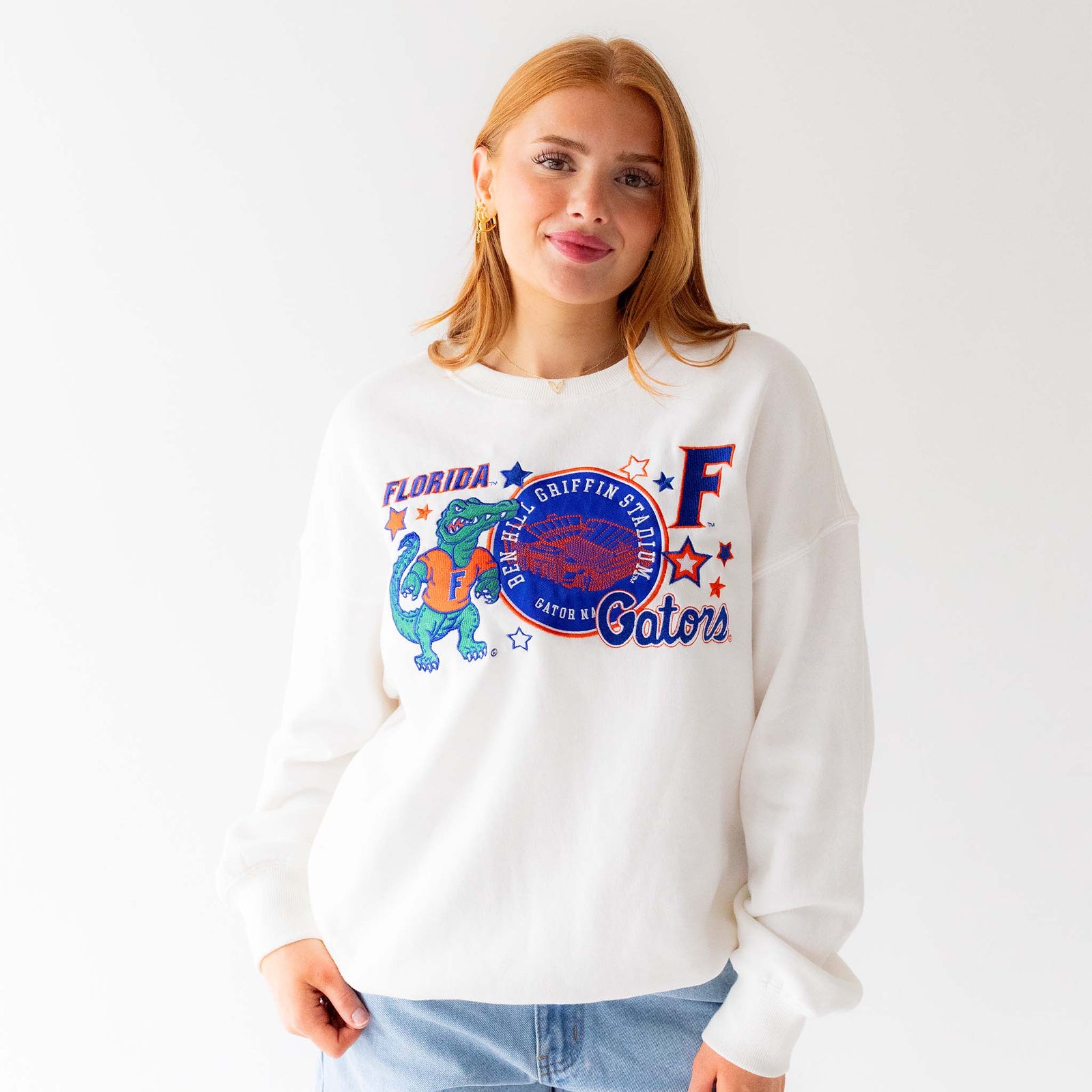 Woman wearing a white crewneck sweatshirt with embroidered Florida Gators graphics including Albert the Alligator mascot, Ben Hill Griffin Stadium patch, letter “F,” and stars.