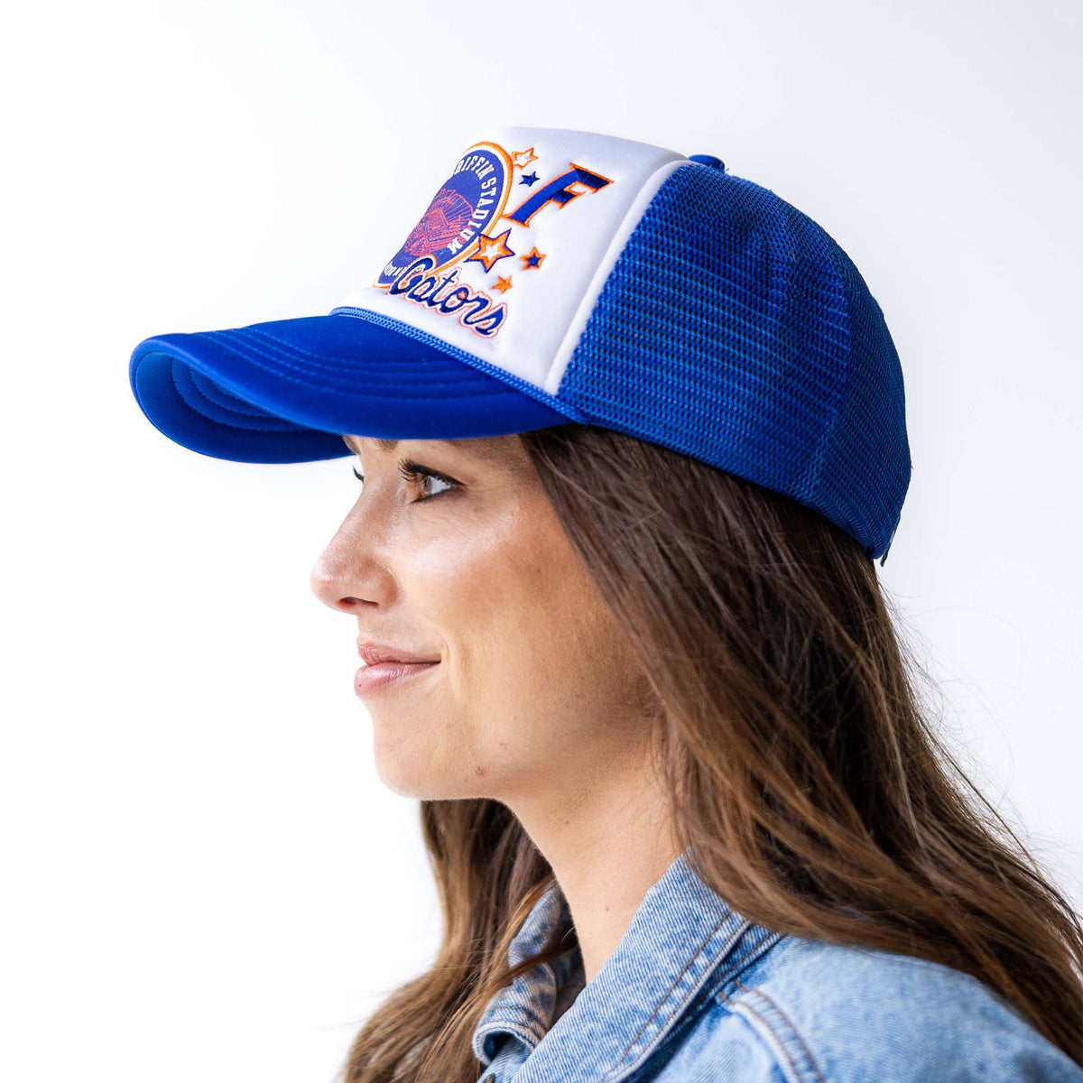 Side view of woman wearing royal blue and white trucker hat with embroidered stadium patch, letter “F,” and stars, showing mesh back and curved brim.