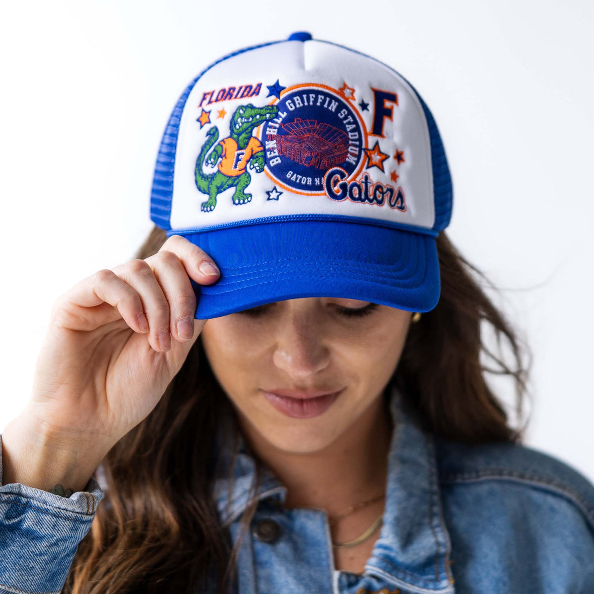 Front view of woman wearing royal blue and white trucker hat with embroidered Florida Gators mascot, Ben Hill Griffin Stadium patch, letter “F,” and stars.