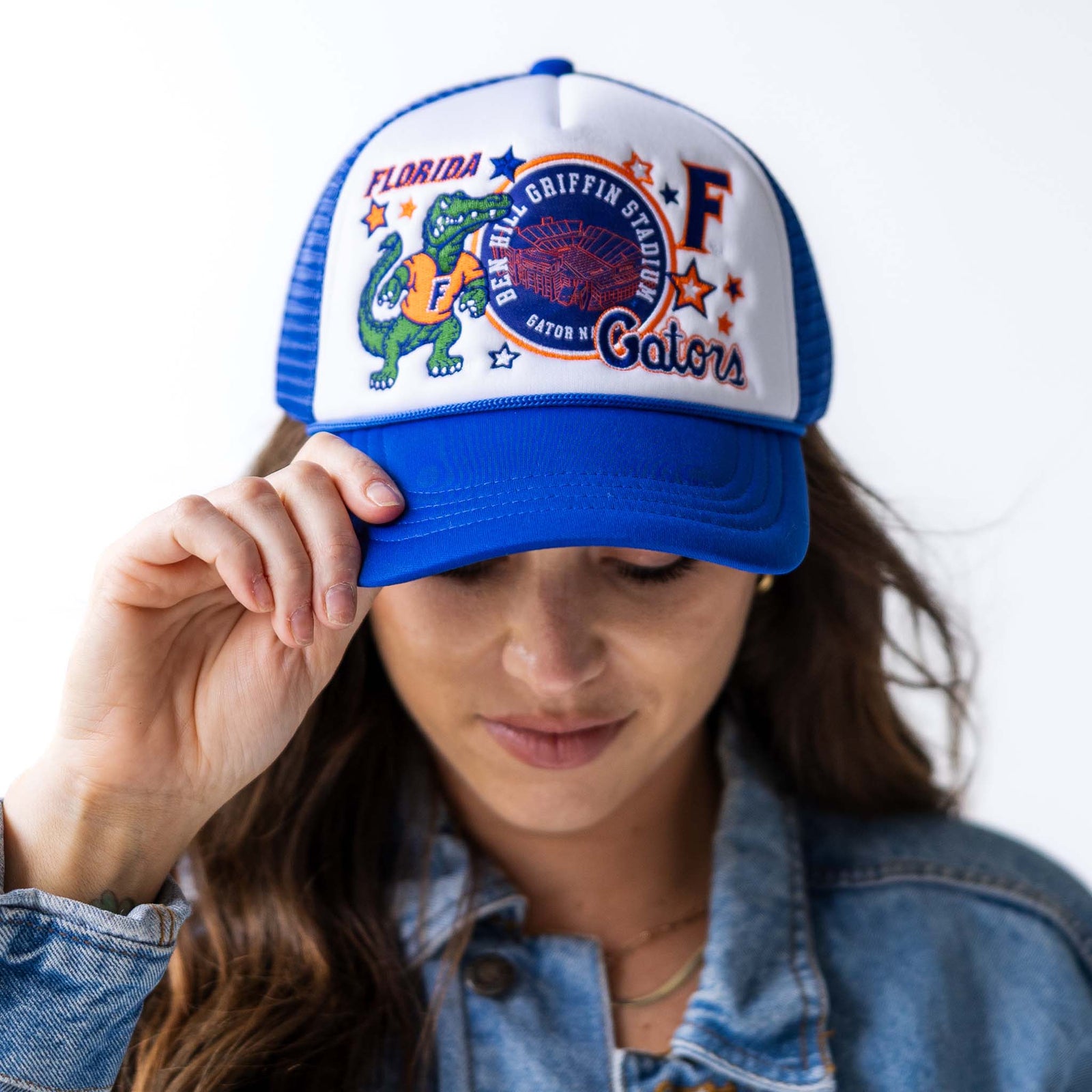 Front view of woman wearing royal blue and white trucker hat with embroidered Florida Gators mascot, Ben Hill Griffin Stadium patch, letter “F,” and stars.