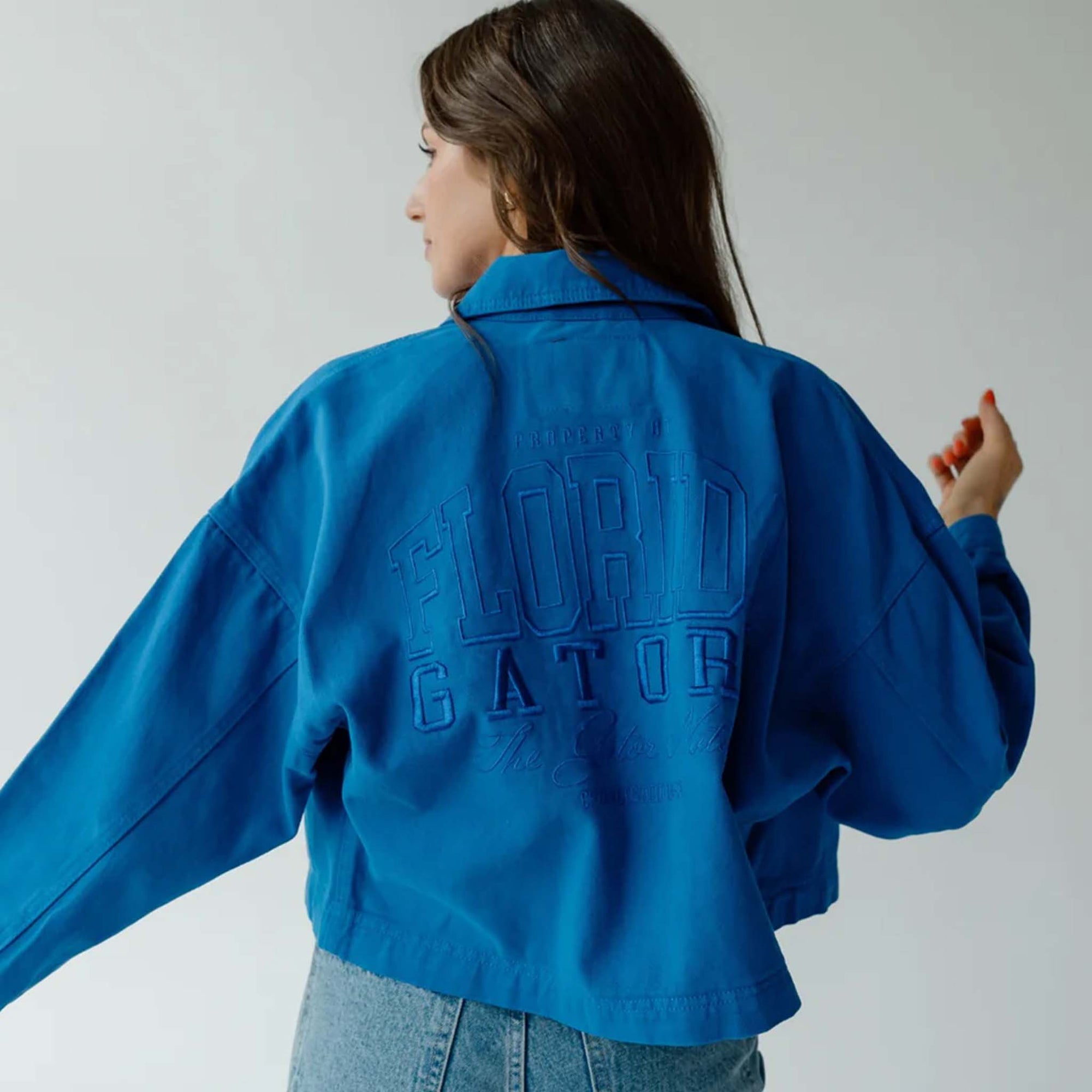Back view of royal blue cropped canvas twill jacket with tonal embroidered text reading “Property of Florida Gators.”