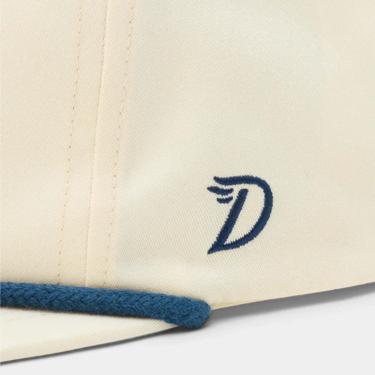 Side detail of ivory Duck Camp hat with navy embroidered “D” logo and blue rope trim along the bill.