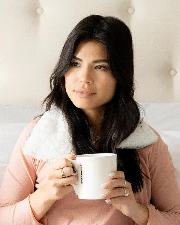 girl sitting in bed drinking coffee and using a warmies neck wrap