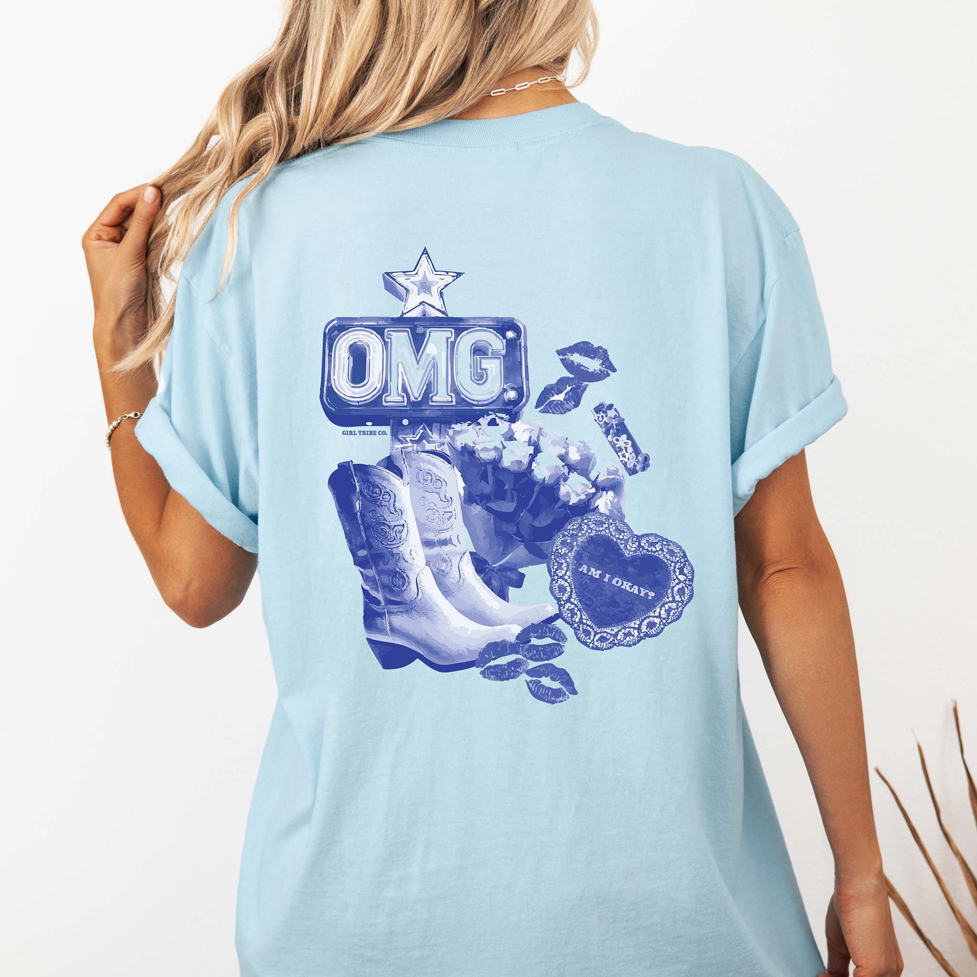 Back view of a woman wearing a light blue t-shirt with rolled sleeves, featuring a large blue graphic of an “OMG” marquee sign, white cowboy boots, roses, lipstick prints, and a lace-trimmed heart with the words “Am I Okay?”