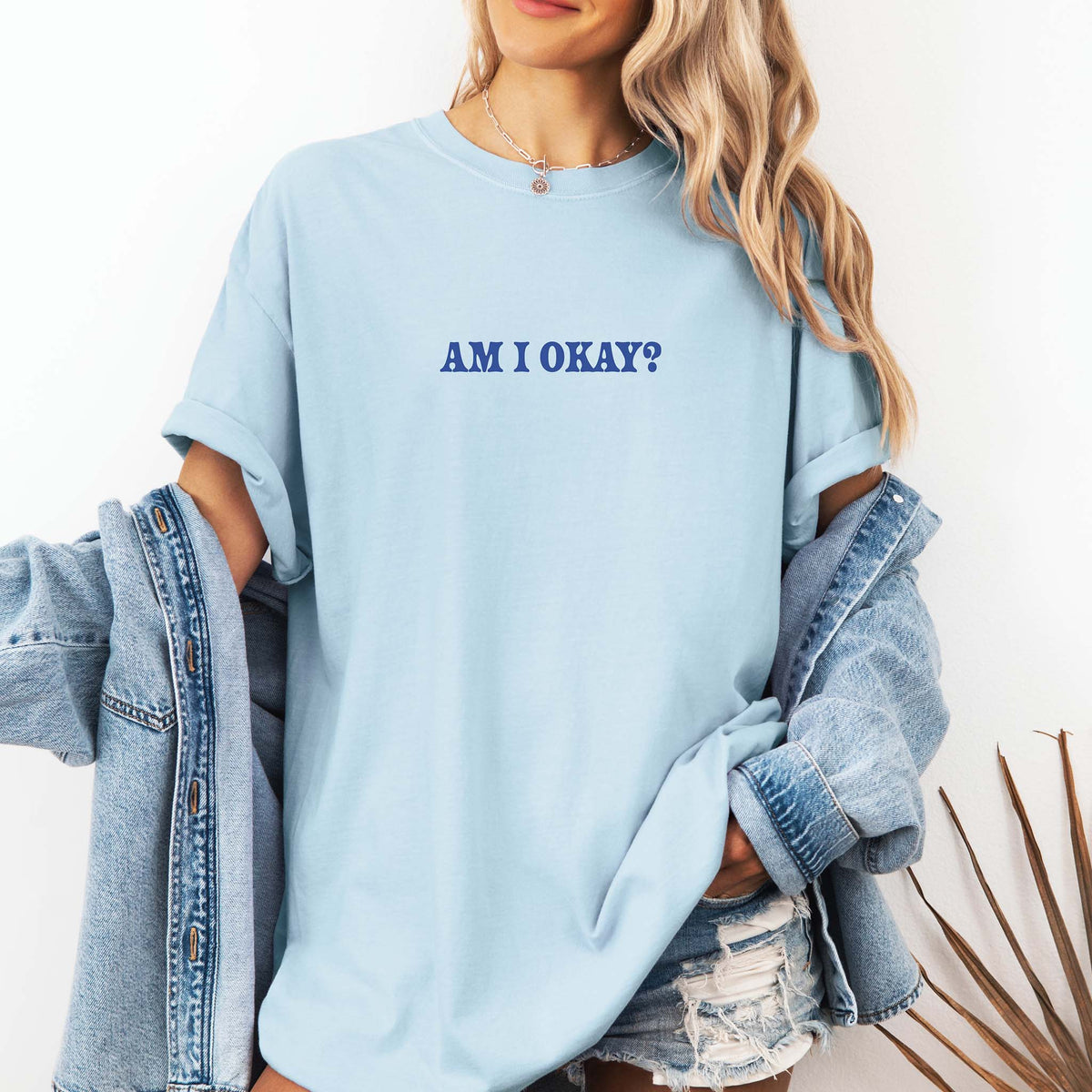 Front view of a woman wearing a light blue t-shirt with rolled sleeves, showing navy text in the center reading “AM I OKAY?” paired with a denim jacket and distressed denim shorts.
