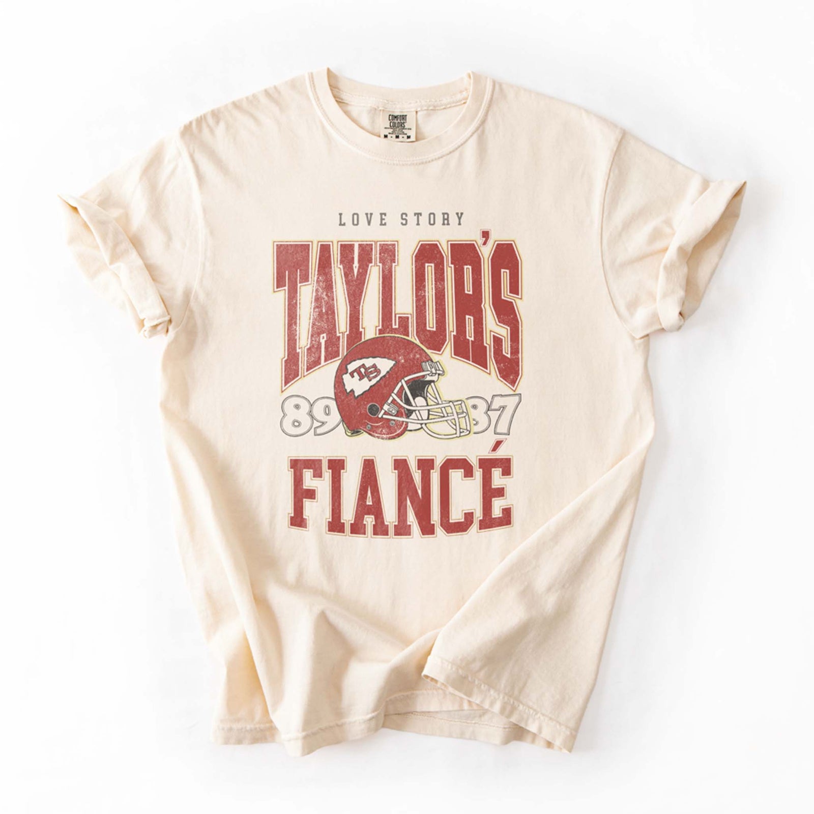 Flat lay of ivory tee with vintage-style football helmet graphic and bold red lettering “Taylor's Fiancé.”