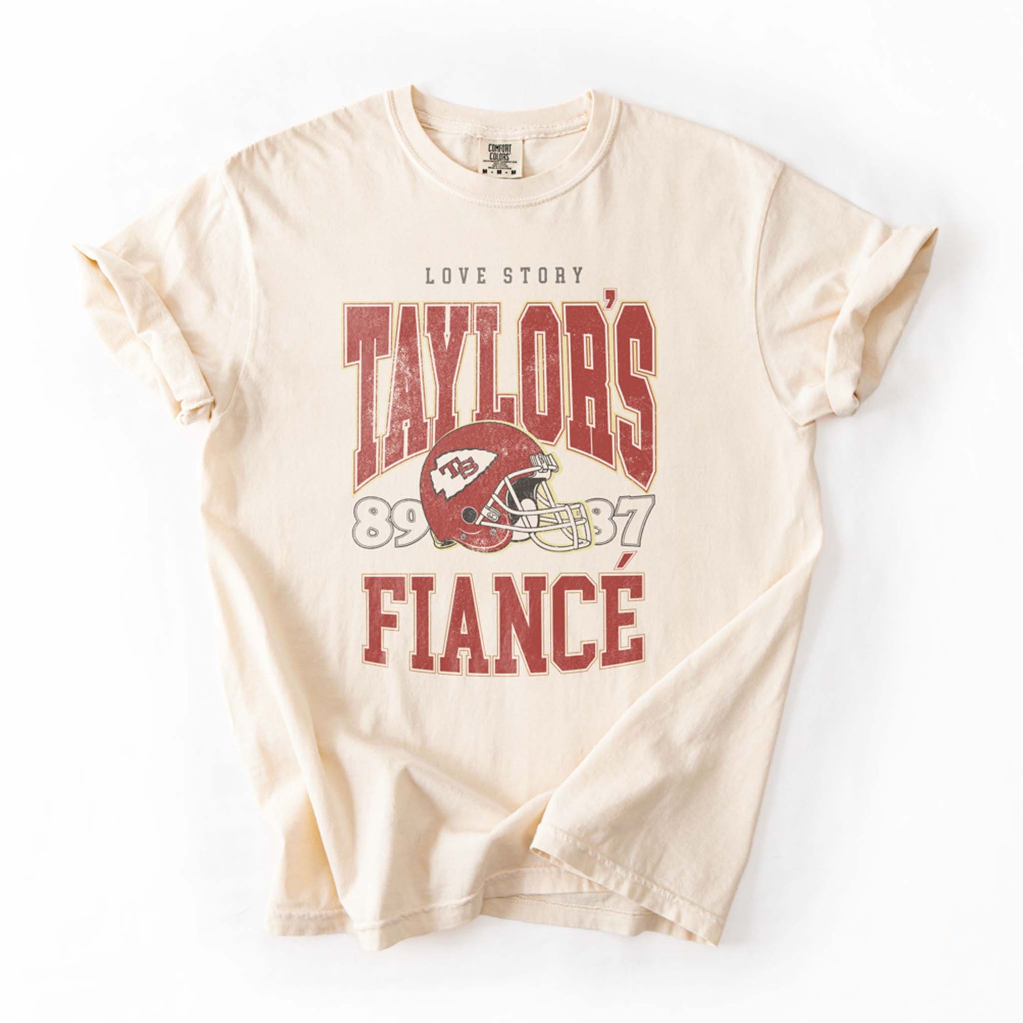 Flat lay of ivory tee with vintage-style football helmet graphic and bold red lettering “Taylor's Fiancé.”