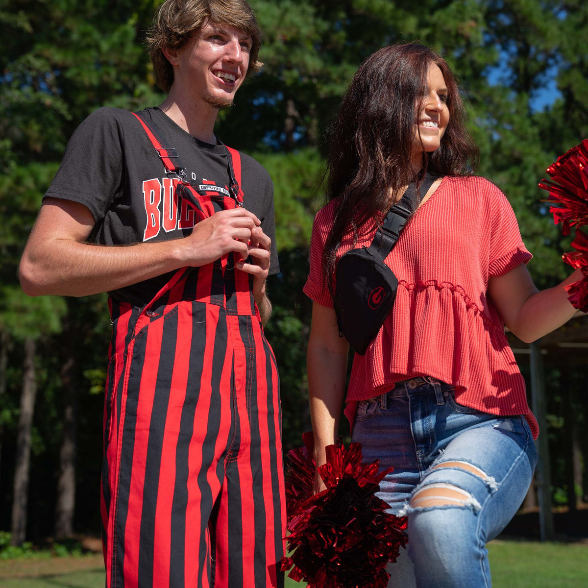 Man wearing red and black striped overalls and “Bulldogs” tee stands with woman in red top holding red pom-poms, both smiling outdoors on a sunny day.