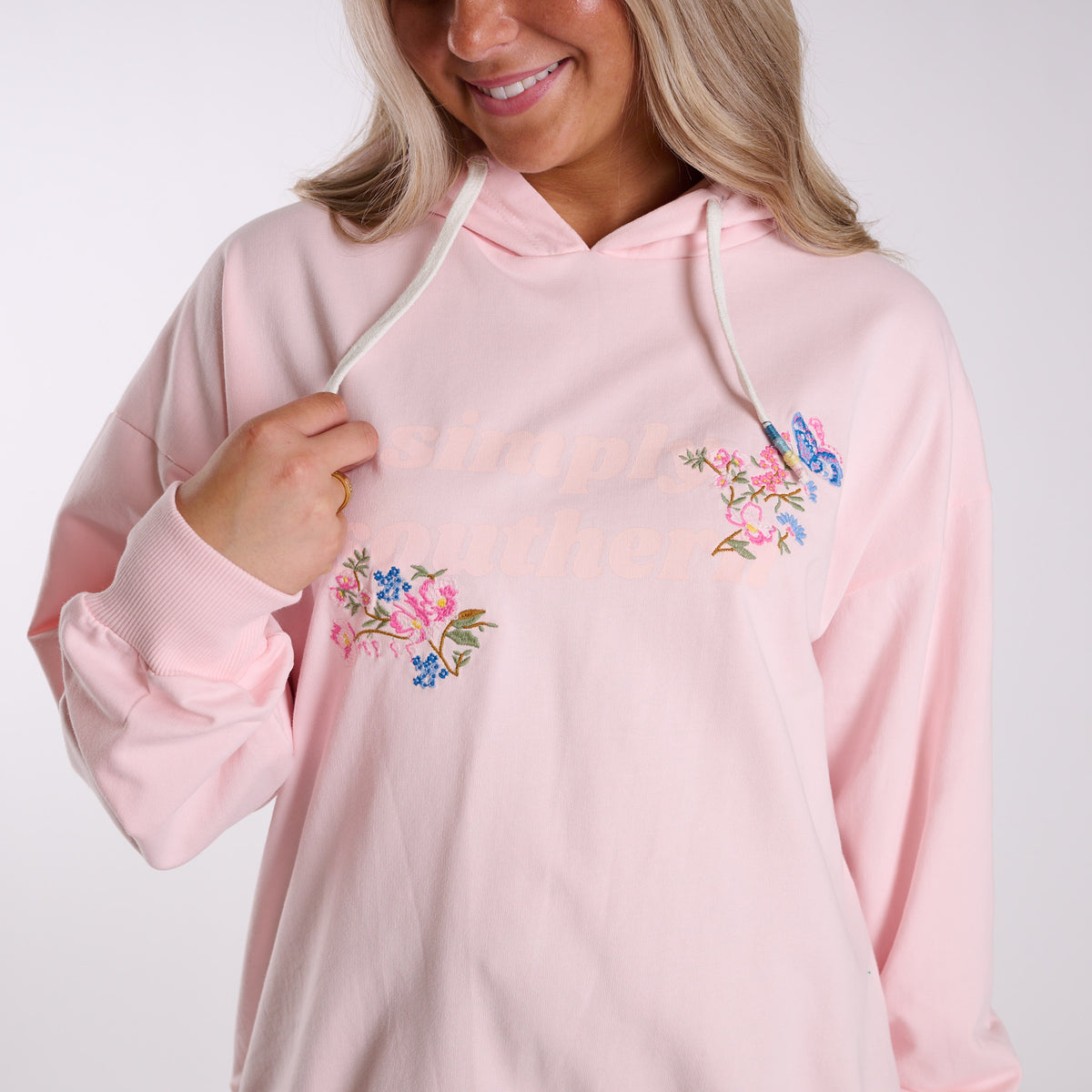 Woman wearing a pink hoodie with floral designs and subtle Simply Southern branding on a white background