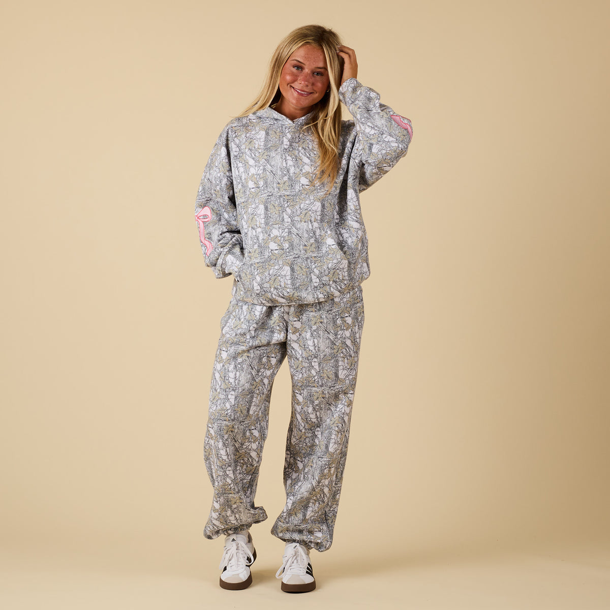Model wearing camo sweatpants with pink bows, paired with matching hoodie.