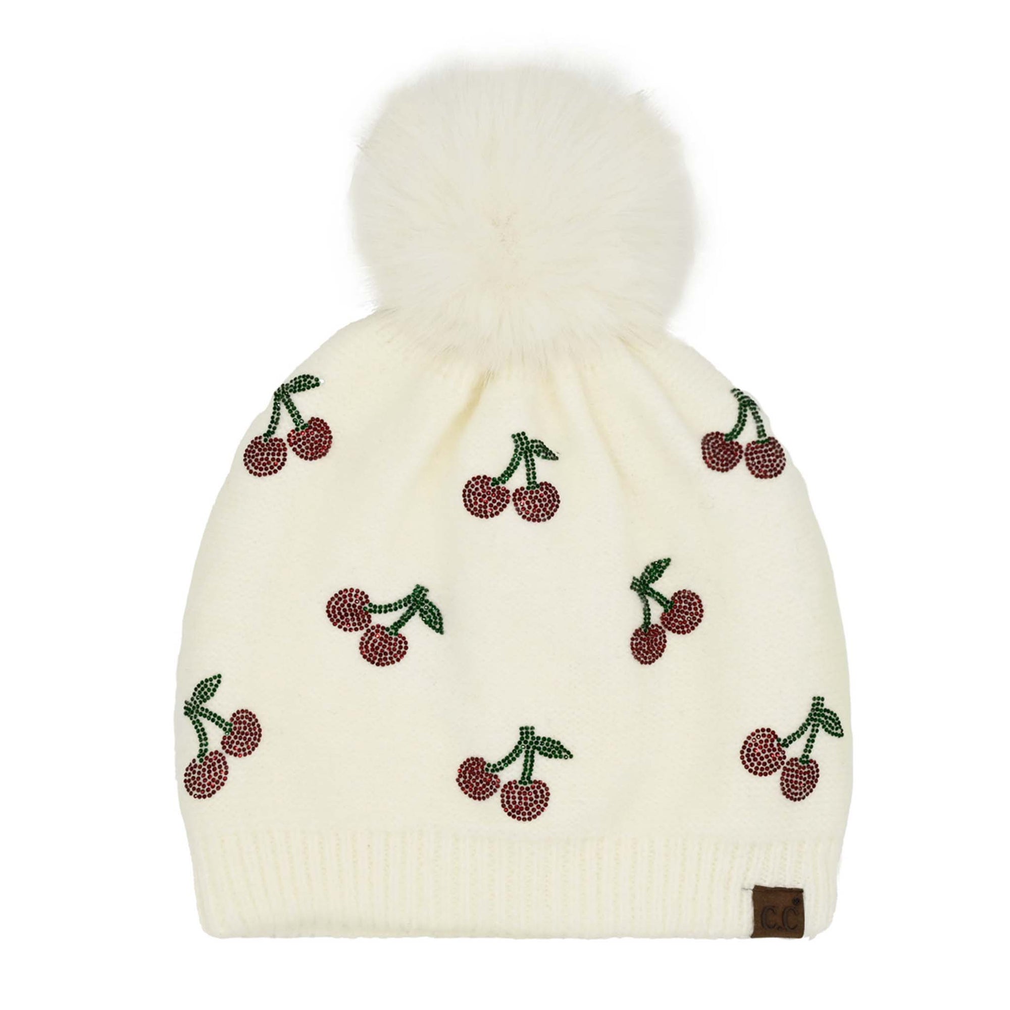 C.C ivory knit beanie with rhinestone cherry embellishments, white faux fur pom on top, and brown leather logo patch on the brim, laid flat on a white background.