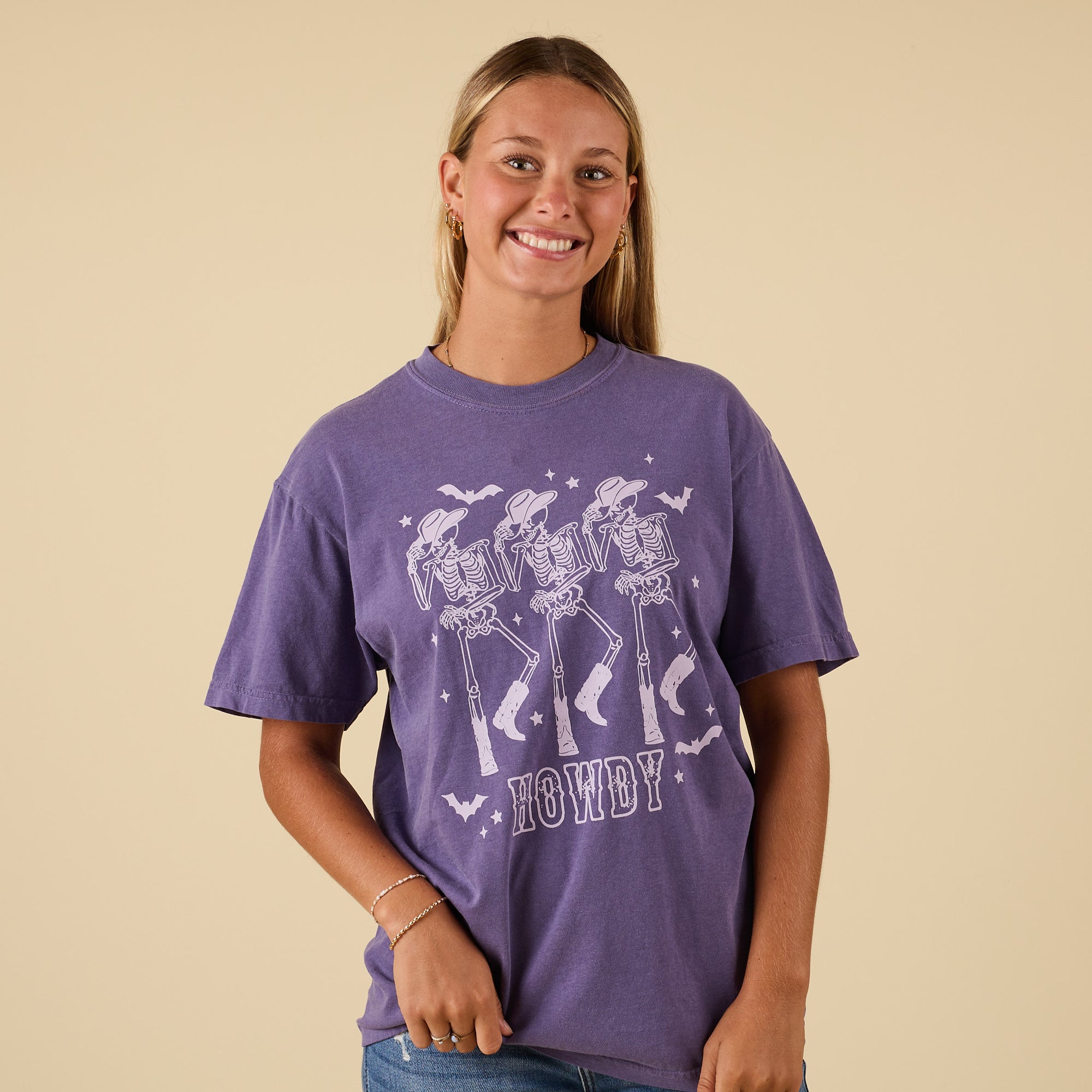 Front view of purple t-shirt featuring dancing skeletons in cowboy hats and boots with ‘Howdy’ text.