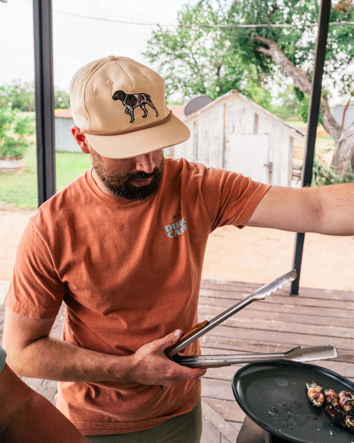 Man wearing tan dog-patch baseball cap and rust graphic T-shirt grilling food on an outdoor porch.