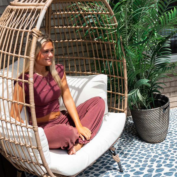 A woman sits cross-legged on a white cushion in a wicker egg chair outdoors. She is wearing a matching maroon lounge set with a cap-sleeve crossover crop top and relaxed pants. A woven basket with a tall green plant sits on a patterned rug next to her, and sunlight filters through the chair's open weave.
