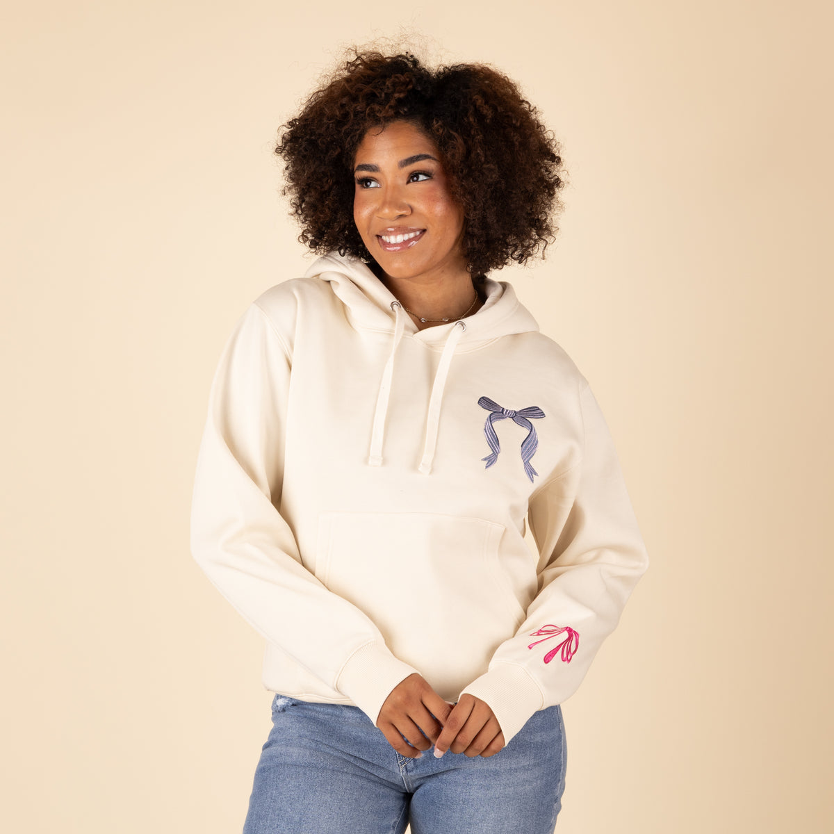 Woman wearing cream-colored hoodie with blue bow graphic on chest and pink bow accent on sleeve, paired with blue jeans
