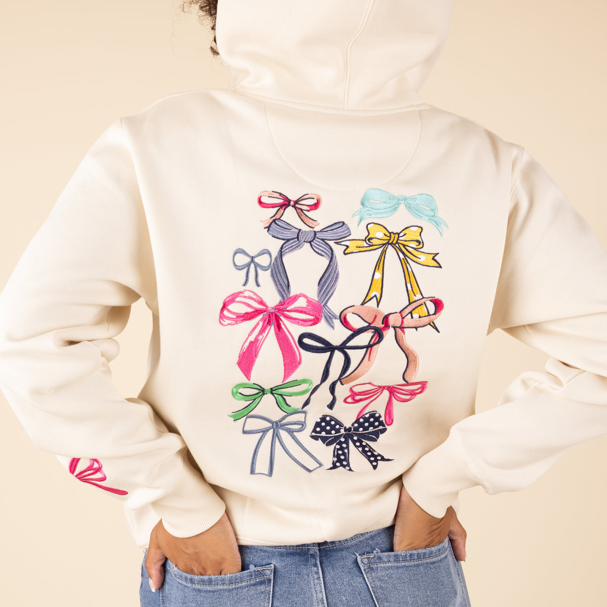 Close-up of cream hoodie showing colorful bow collection graphic with various ribbons and bows in pink, blue, yellow, green, and navy colors across the back