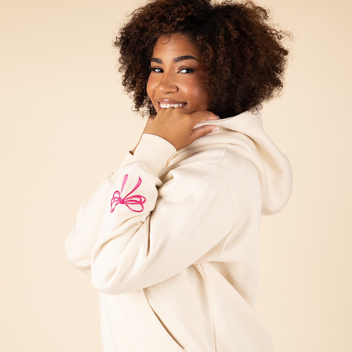 Woman in cream hoodie with pink bow detail on sleeve cuff, hands positioned near face in casual pose