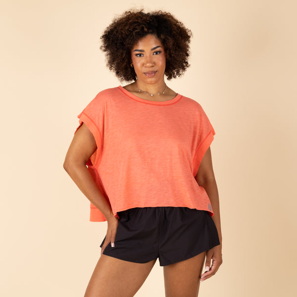 Free People Movement My Time Tee in Neon Coral, sizes XS–XL. Full front view of model wearing the relaxed-fit tee, hands in shorts pockets, showcasing the draped silhouette.