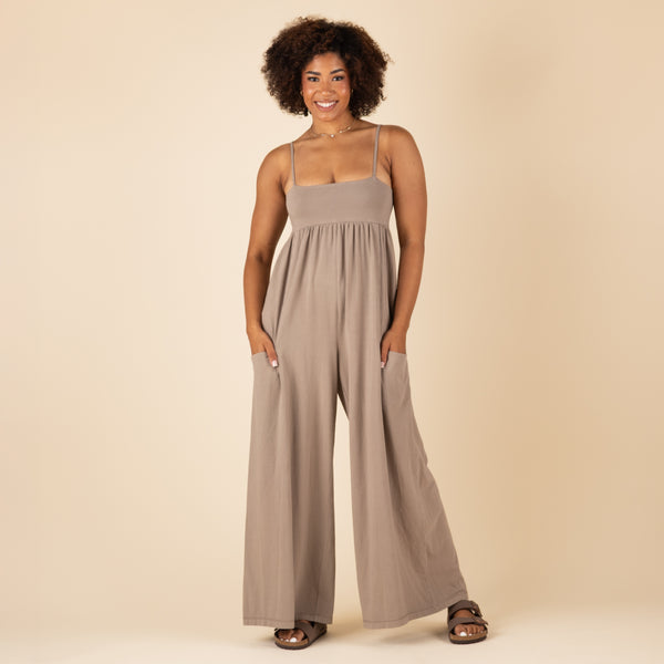  Woman wearing a taupe wide-leg jumpsuit with spaghetti straps and pockets, standing front-facing with hands in pockets against a beige background.