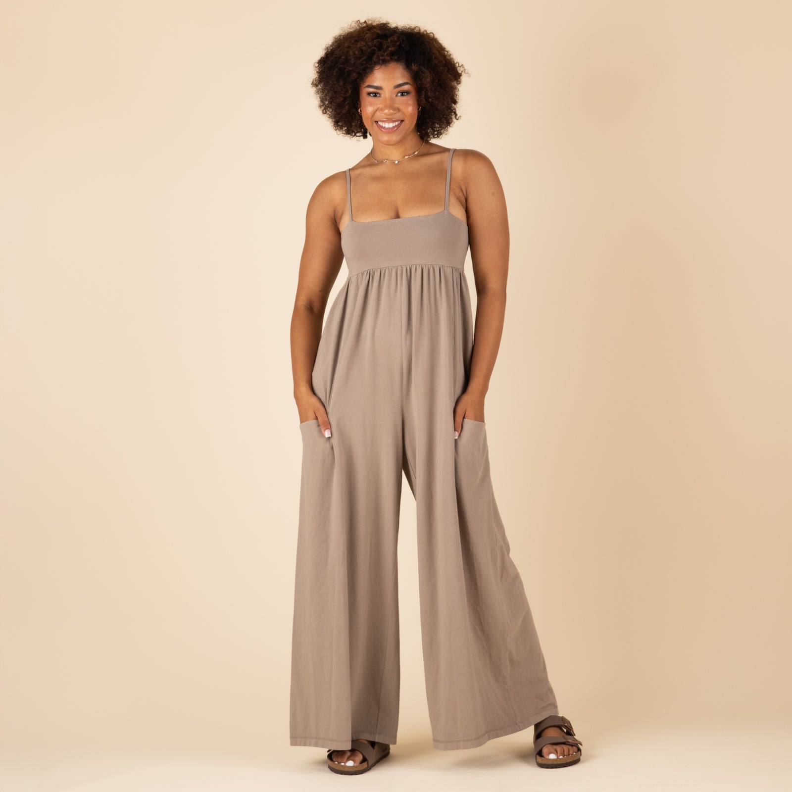 Woman wearing a taupe wide-leg jumpsuit with spaghetti straps and pockets, standing front-facing with hands in pockets against a beige background.