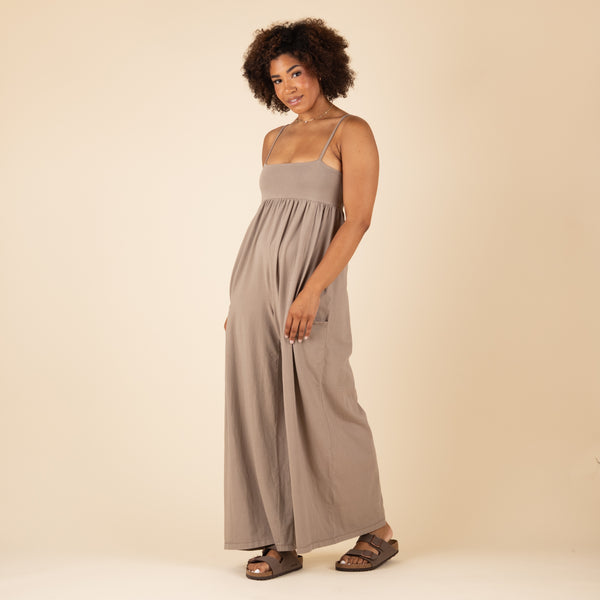  Side view of a woman in a taupe spaghetti strap jumpsuit with a relaxed fit and flowy wide legs, styled with brown sandals.