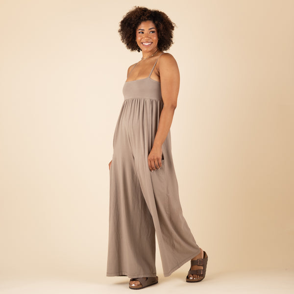  Angled side view of a woman wearing a taupe wide-leg jumpsuit with spaghetti straps and pockets, smiling and walking slightly forward.
