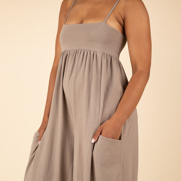  Close-up detail of the taupe jumpsuit showing the gathered empire waist, spaghetti straps, and hand in a front pocket.