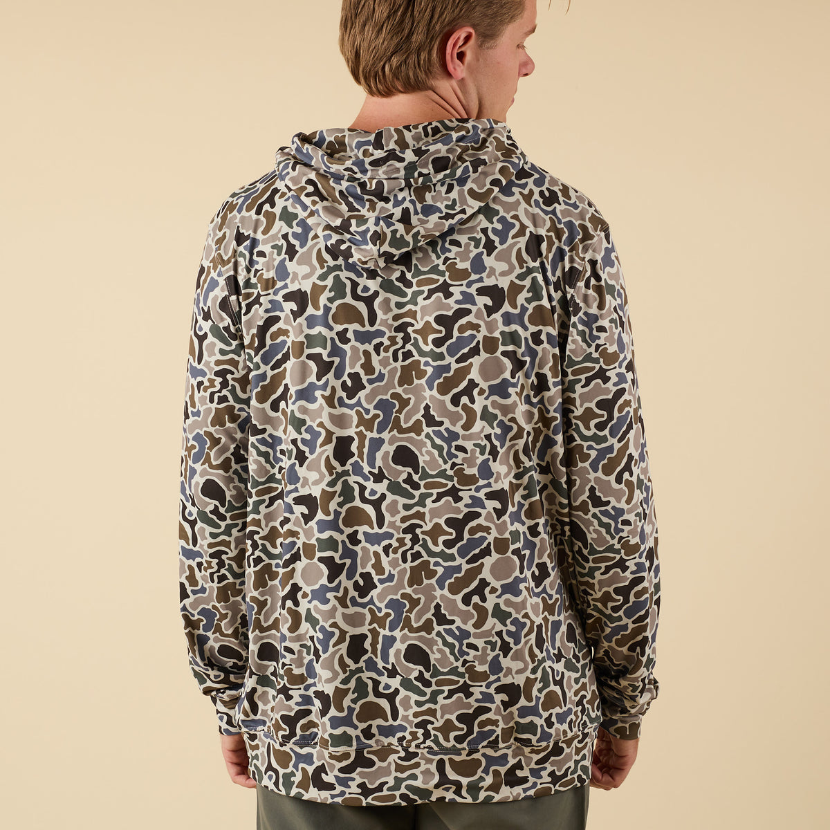 Back view of a man wearing a camo performance hoodie on a beige background.