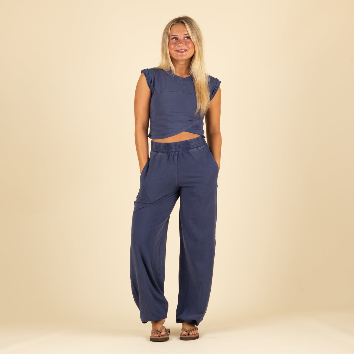 Front view of woman in navy twist-front crop top and wide-leg joggers with elastic waist and cuffs, smiling with hands in pockets.
