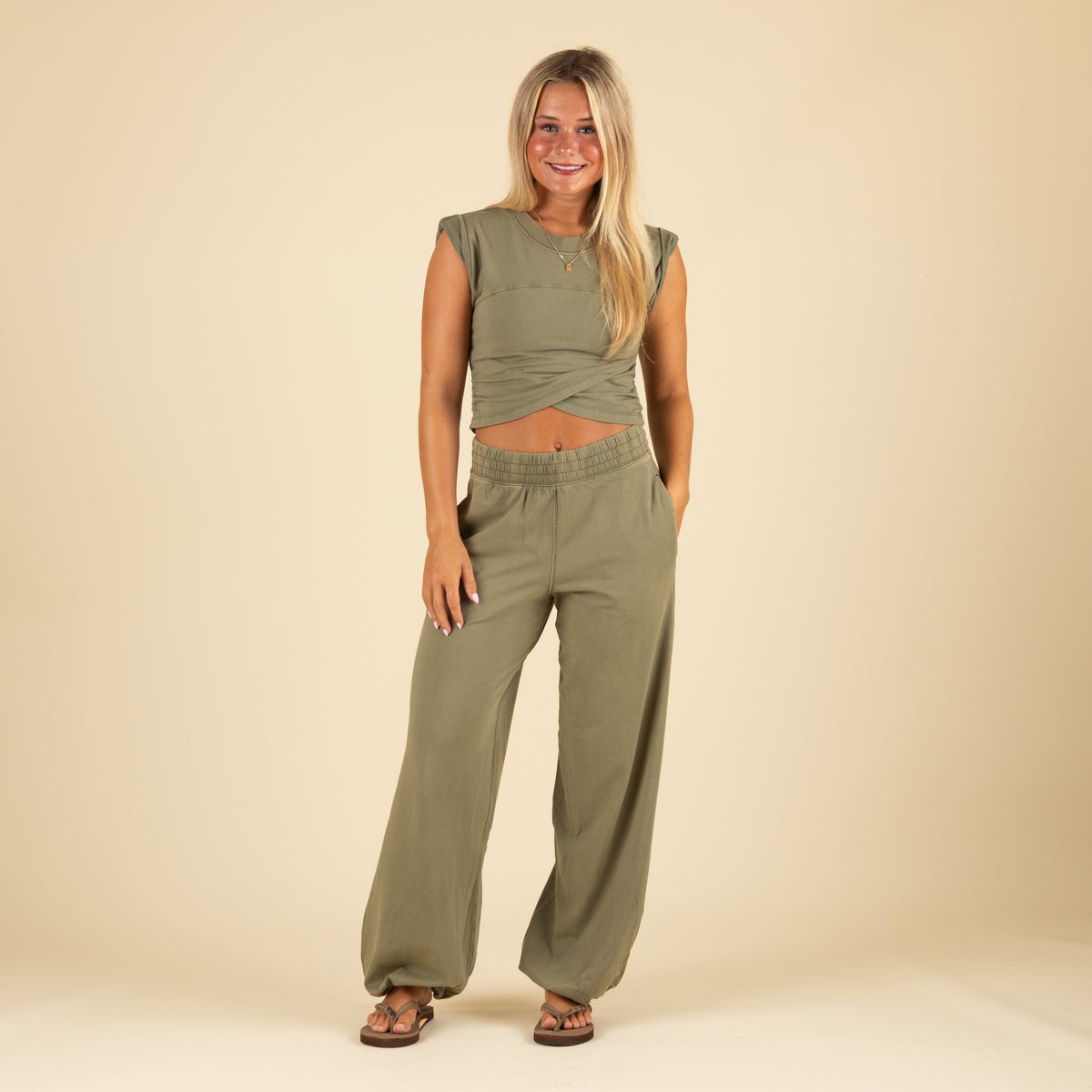 Full front view of woman in olive green twist-front crop top and wide-leg joggers with elastic cuffs and side pockets, smiling with hands in pockets.