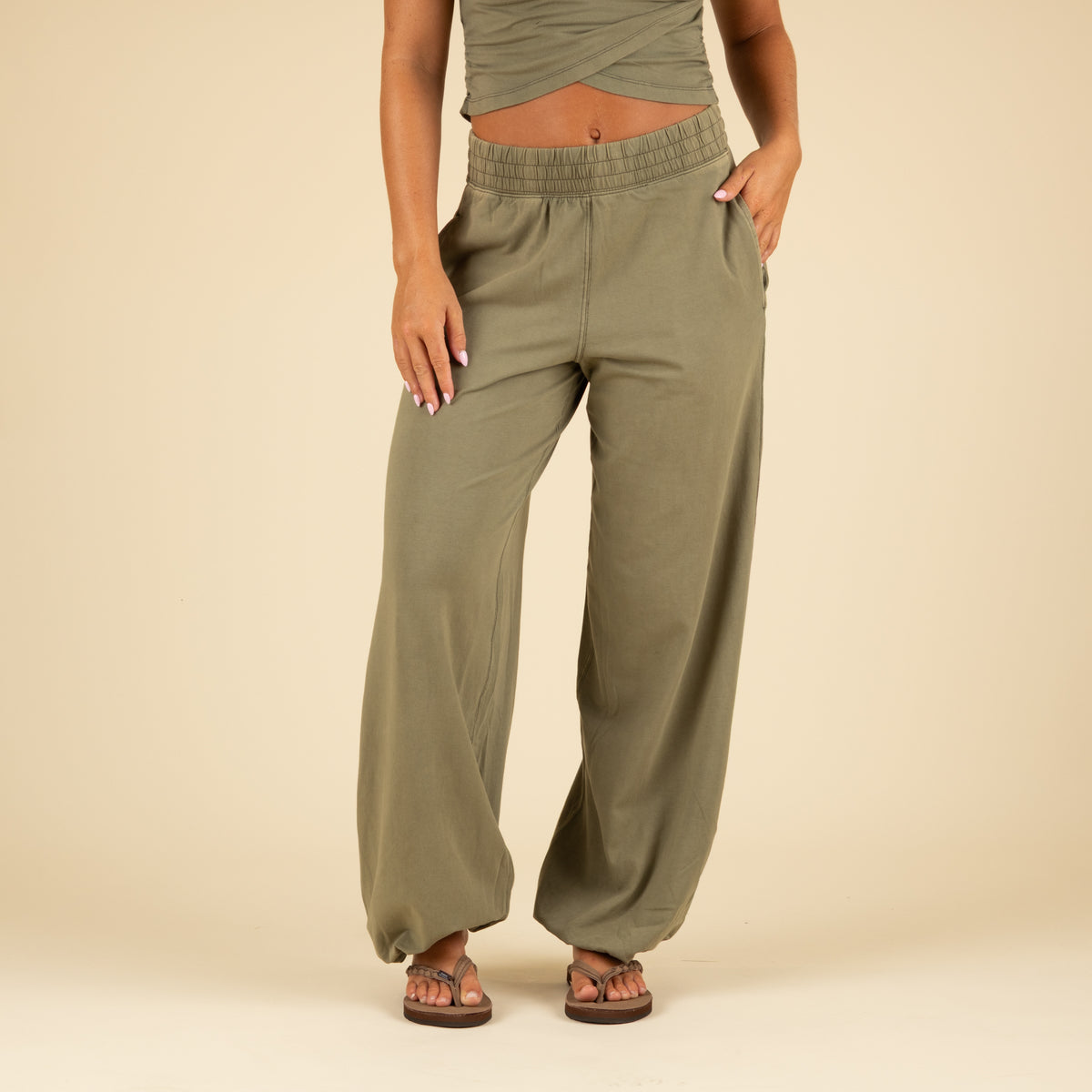 Focus on lower half of olive green jogger pants with wide legs, elastic cuffs, and side pockets, paired with brown sandals.