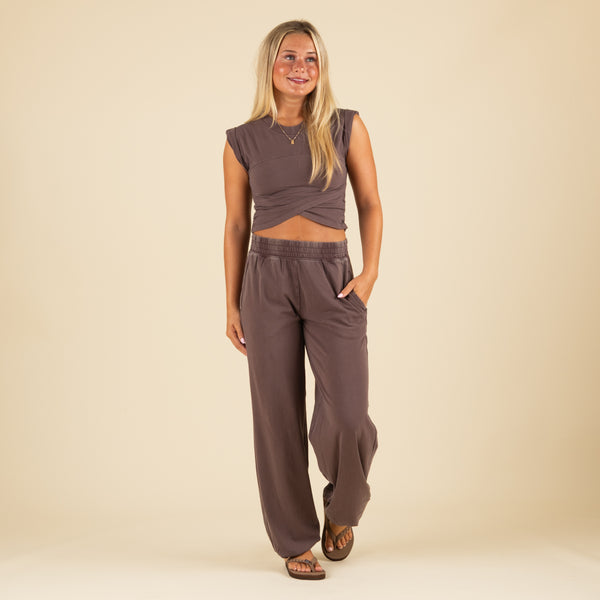  Front view of woman wearing brown wrap crop top and wide-leg pants with side pockets, one hand in pocket, smiling.