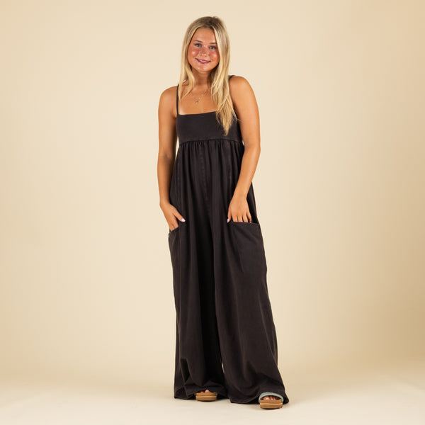  Woman wearing a black wide-leg jumpsuit with spaghetti straps and front pockets, standing and smiling with hands in pockets against a beige background.