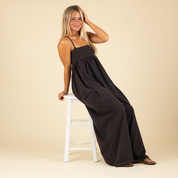  Woman sitting on a white stool in a black wide-leg jumpsuit with spaghetti straps, posing with one hand on her head and smiling.
