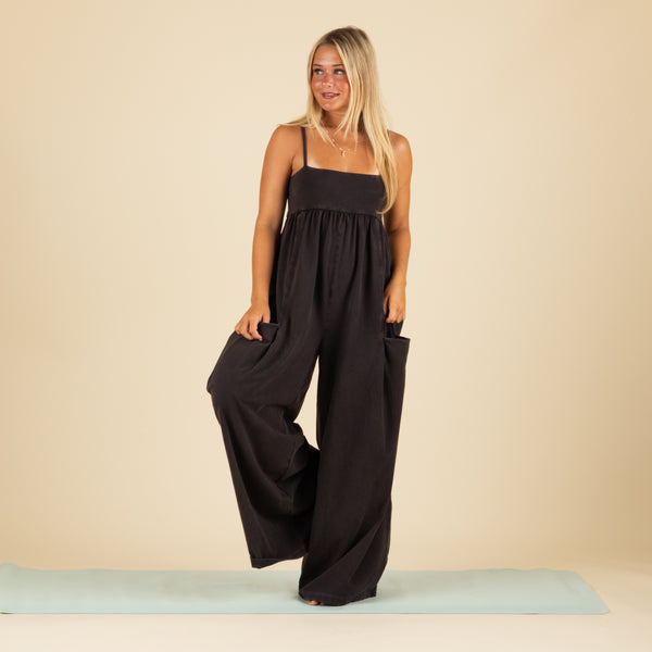  Woman balancing on one leg in a black wide-leg jumpsuit with spaghetti straps and front pockets, standing barefoot on a light blue yoga mat.