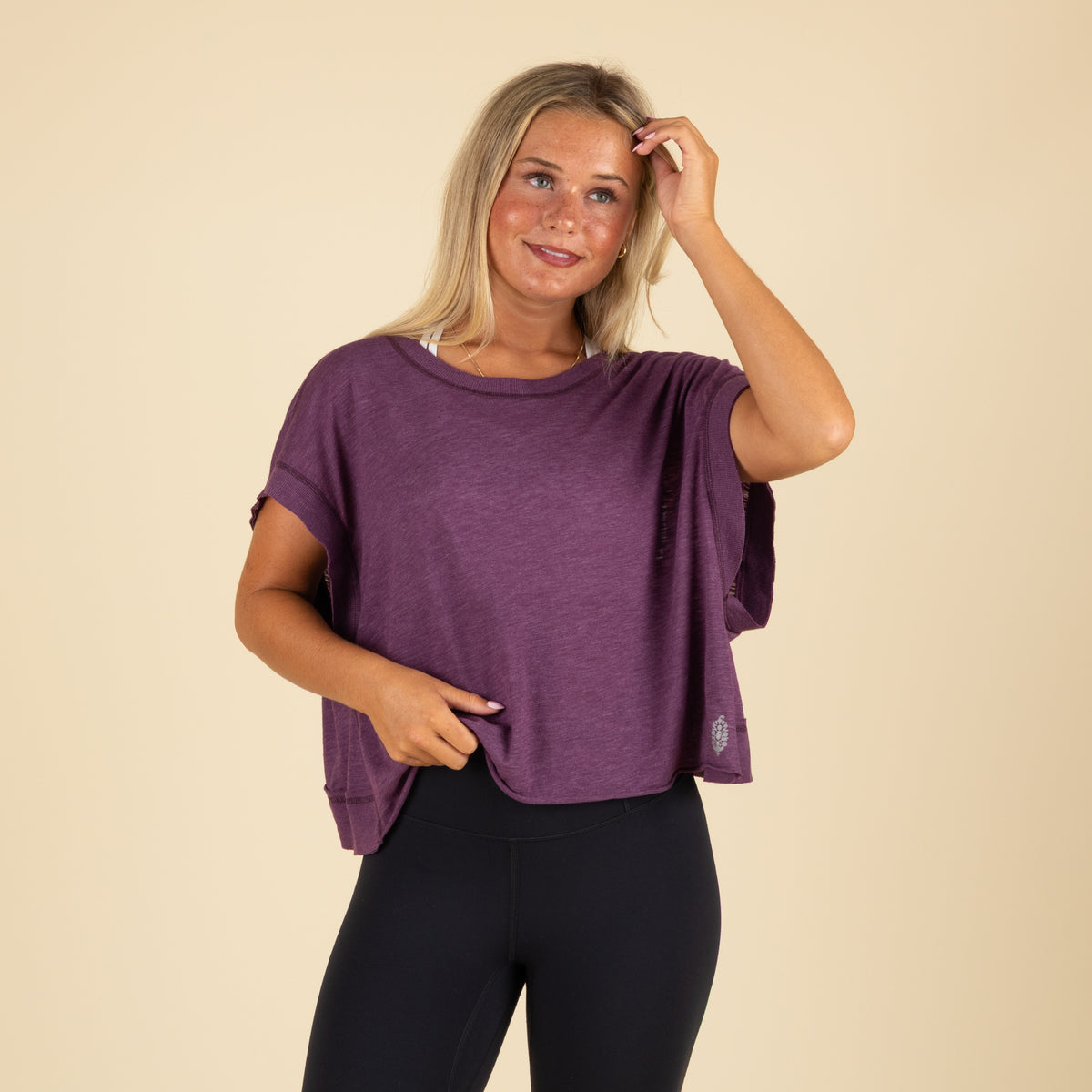 Woman smiling in a purple short sleeve crop top and black leggings, gently lifting the front hem of the top.