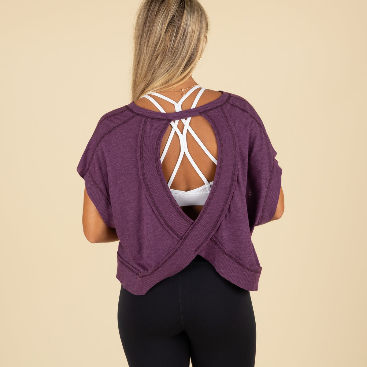 Back view of a woman wearing a purple cropped top with a crossover open back over a white strappy sports bra and black leggings.