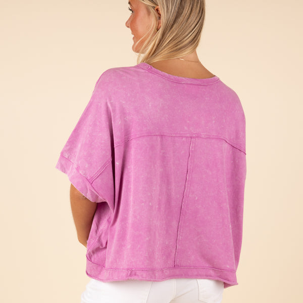  Very J Boxy Pocket Short Sleeve Crop Top, sizes S-L, color orchid. Woman facing away from camera in an orchid purple cropped t-shirt with a relaxed fit and front pocket, paired with white pants.