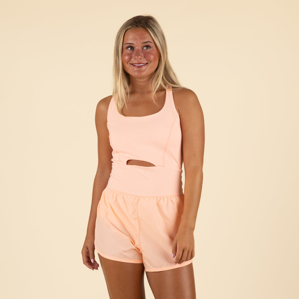 Woman wearing a soft peach/coral colored athletic romper with similar cutout waist design, standing naturally with arms at sides, smiling at camera against beige background