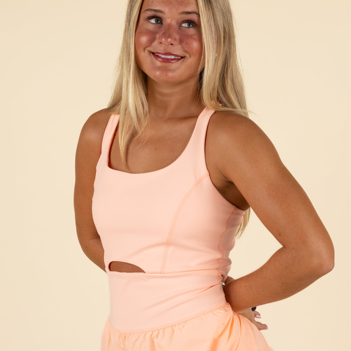 Close-up front view of woman in peach athletic romper with hand on hip, highlighting the cutout waist detail and scoop neckline of the garment