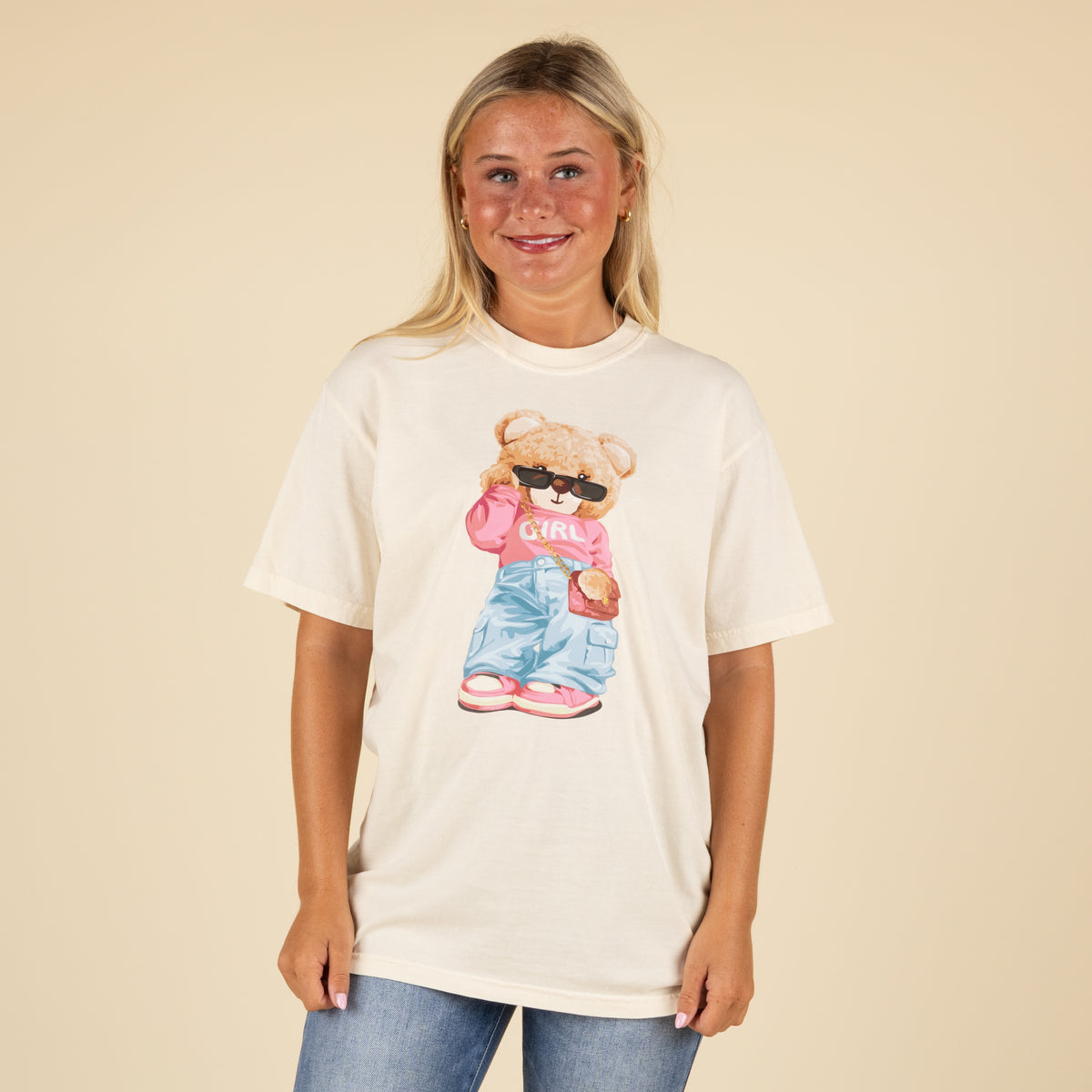 Cream t-shirt with graphic of a teddy bear wearing sunglasses, pink “GIRL” top, cargo jeans, pink sneakers, and a purse, shown on a smiling woman in jeans.