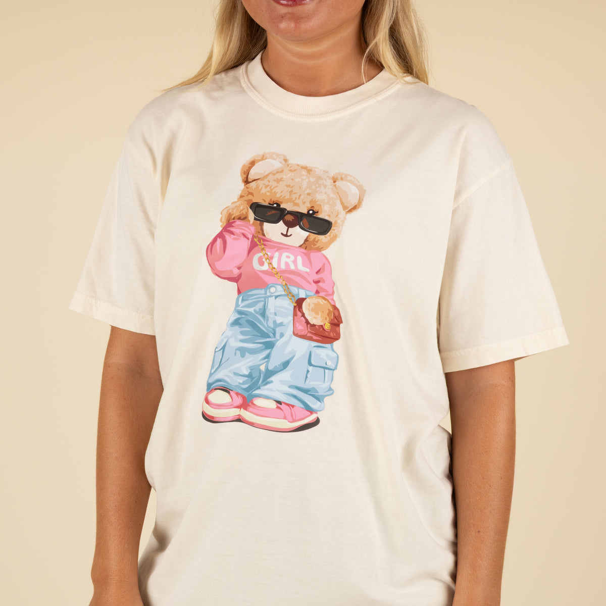 Cream t-shirt with graphic of a teddy bear wearing sunglasses, pink “GIRL” top, cargo jeans, pink sneakers, and a purse.