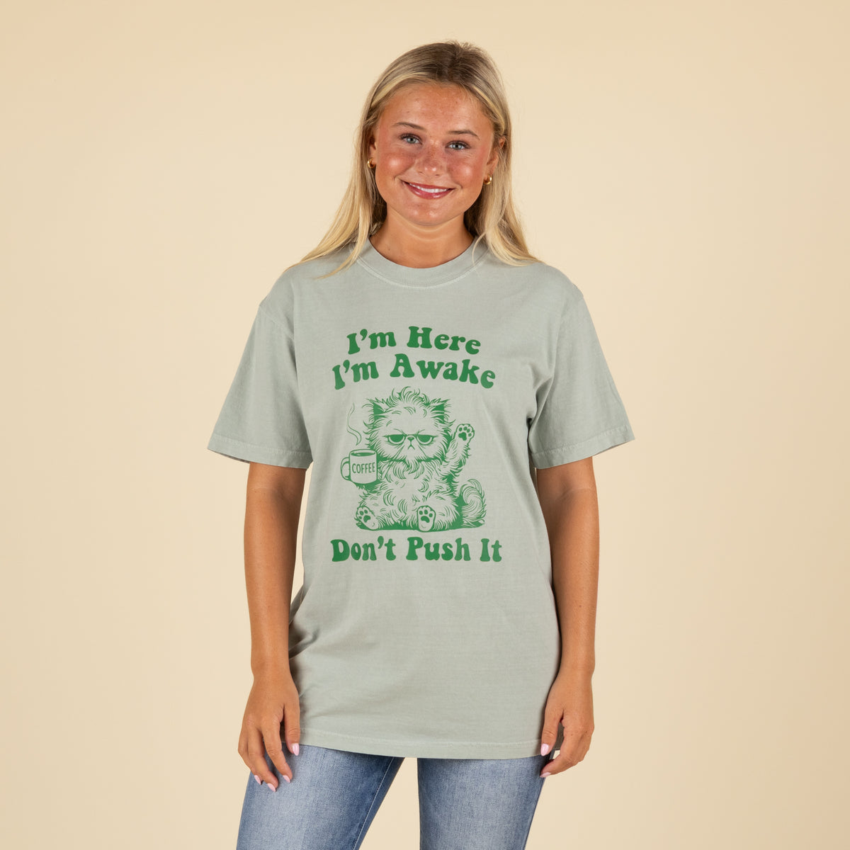 Light sage green t-shirt with green graphic of a grumpy cat holding a coffee mug and text that reads “I’m Here I’m Awake Don’t Push It,” worn by a smiling woman in jeans.