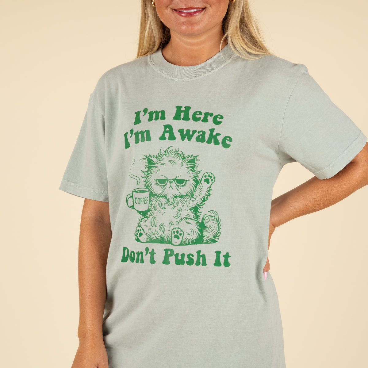 Light sage green t-shirt with green graphic of a grumpy cat holding a coffee mug and text that reads “I’m Here I’m Awake Don’t Push It,” worn by a smiling woman in jeans.
