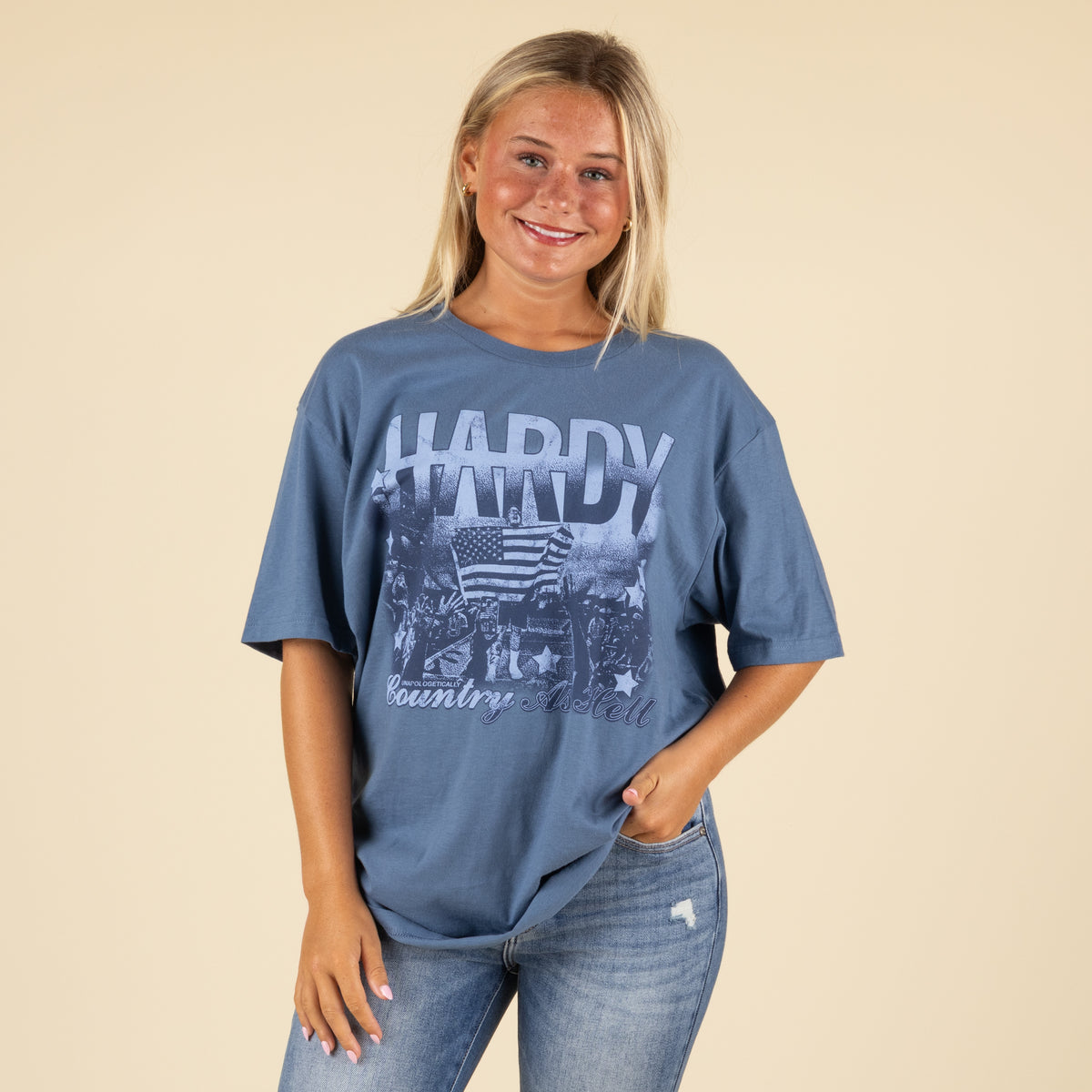 Woman smiling in blue oversized Hardy “Country As Hell” graphic t-shirt with her hand in her jeans pocket.