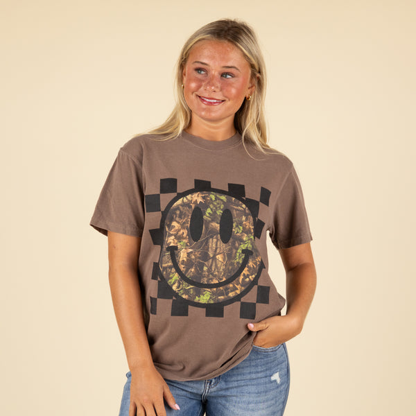 Person wearing a brown t-shirt with a camouflage smiley face design on a beige background