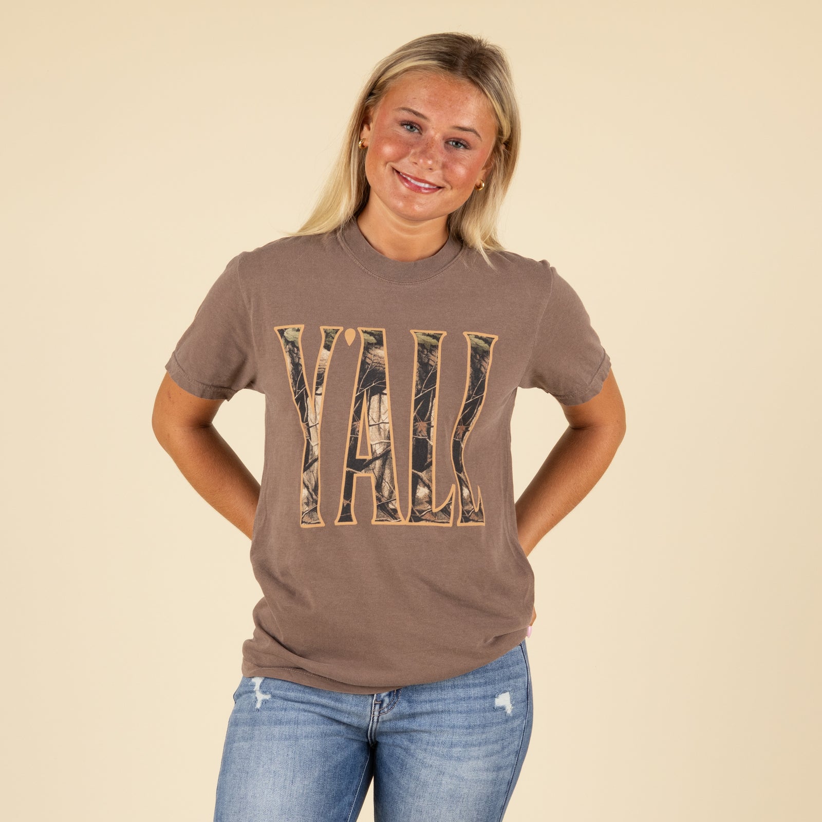 Person wearing a brown t-shirt with 'Yall' printed in camouflage style on a beige background