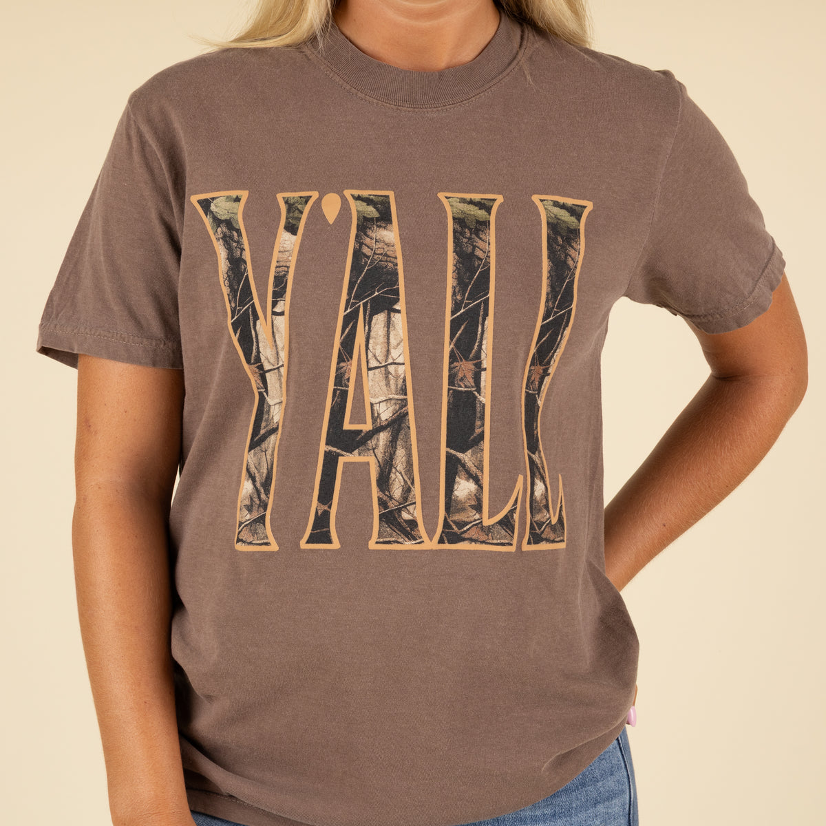 Person wearing a brown t-shirt with 'Y'all' in camouflage print on a beige background