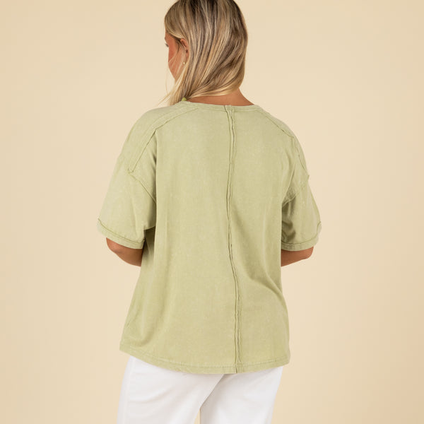 Back view of a woman wearing a sage green oversized t-shirt with visible back seam detail and rolled sleeves.