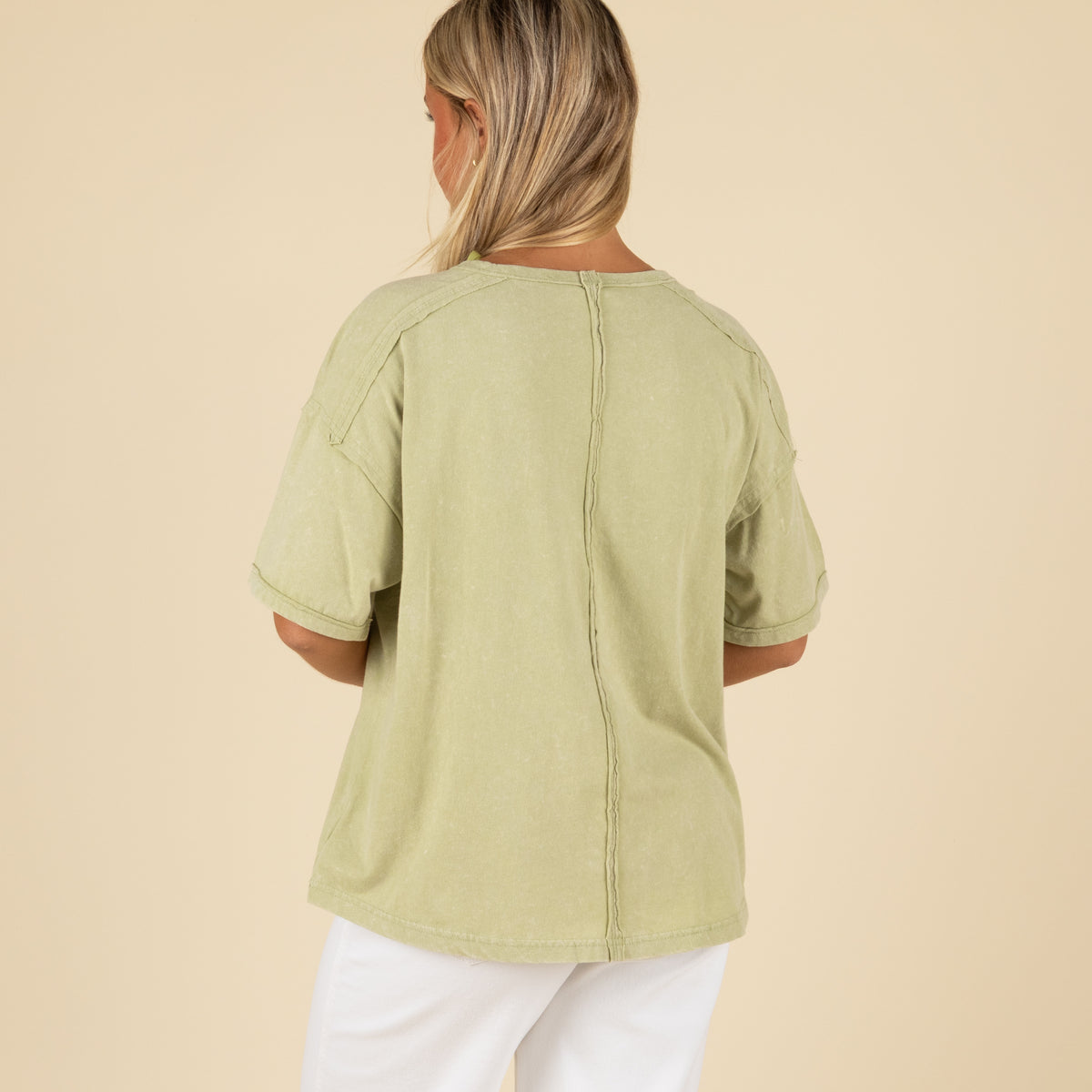 Back view of a woman wearing a sage green oversized t-shirt with visible back seam detail and rolled sleeves.